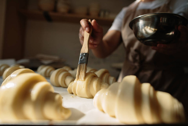 Female baker smears raw croissants with a brush in the yolk, the cooking process. - Photo, image