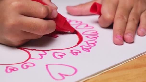 Close-up van een klein meisje dat een hart tekent en kleurt met haar gekleurde stiften. Schattig jong meisje doet huiswerk aan tafel thuis. Kunstnijverheid. - Video