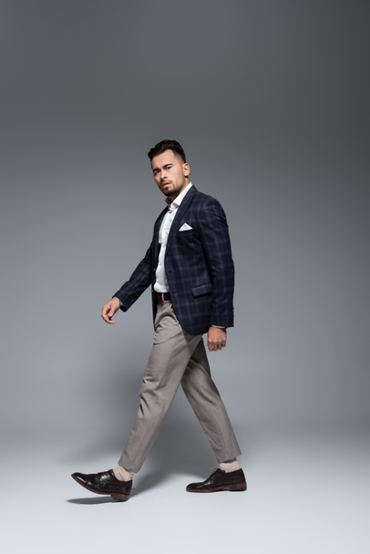 longitud completa de joven hombre de negocios barbudo en traje caminando sobre gris - Foto, Imagen