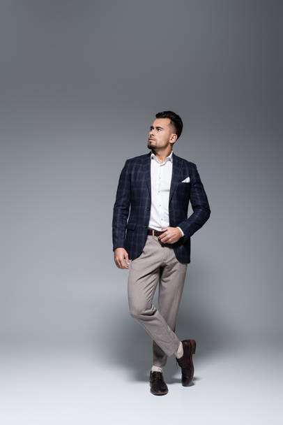 full length of young bearded businessman in suit standing with crossed legs on grey - Foto, imagen
