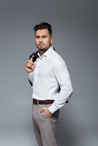 young bearded businessman holding blazer and standing with hand in pocket on grey - Foto, imagen