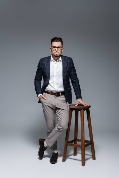 homme d'affaires barbu en costume et lunettes debout avec la main dans la poche près de la chaise sur gris - Photo, image
