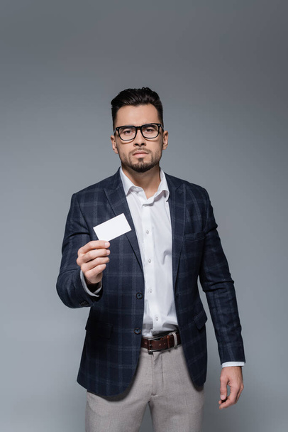 joven hombre de negocios en gafas y chaqueta a cuadros que sostiene la tarjeta en blanco aislado en gris - Foto, imagen
