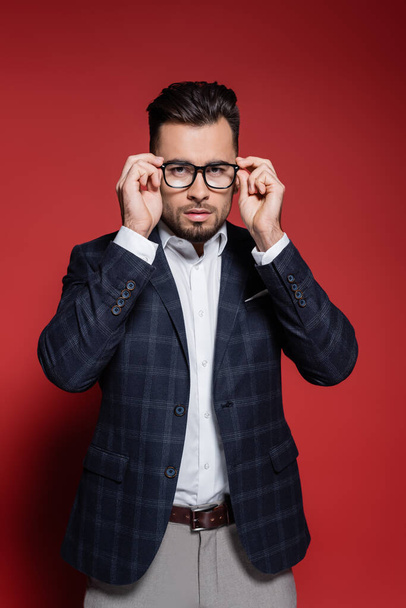 young businessman in plaid blazer adjusting glasses on red - Фото, изображение