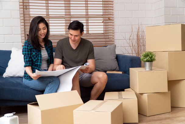 Smiling young Asian happy couple hold blueprint for home decoration ideas at moving day in their new home after buying real estate. Concept of starting a new life for a newly married couple. - Photo, Image