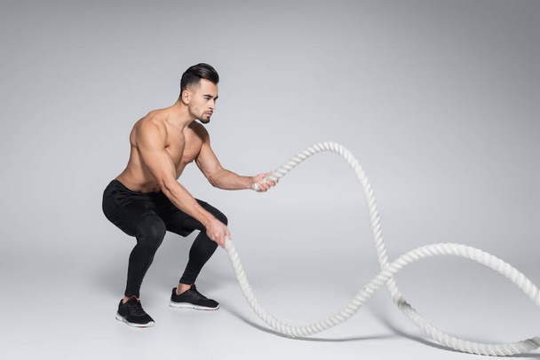 full length of muscular sportsman exercising with battle ropes on grey - Foto, Imagem