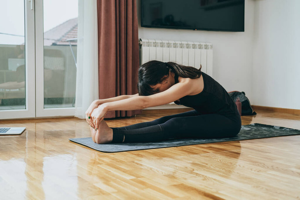 An Anonymous Woman Doing Warm Up Exercises. Unrecognizable fitness woman stretching arms and legs while sitting in paschimottanasana pose on a gray exercise mat in the living room at home. - Foto, imagen
