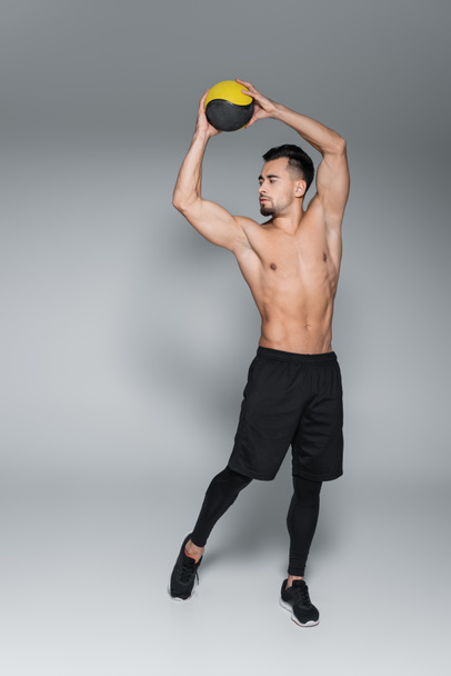 full length of shirtless and bearded sportsman holding ball above head on grey - Foto, imagen