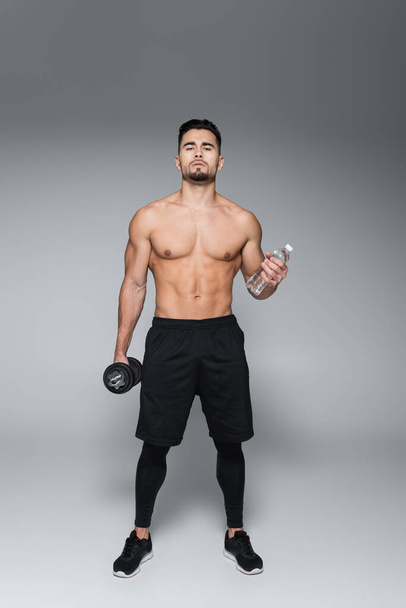 full length of shirtless sportsman holding dumbbell and bottle with water on grey - Фото, изображение