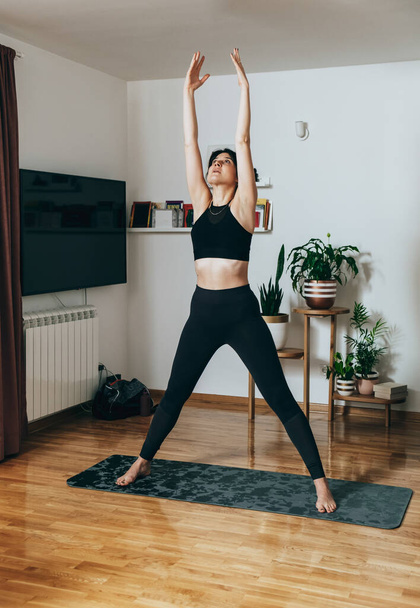Hermosa mujer fitness en ropa deportiva negra estirando brazos y hombros y haciendo ejercicios de respiración mientras está de pie en una alfombra de ejercicio gris en la sala de estar en casa - Foto, imagen