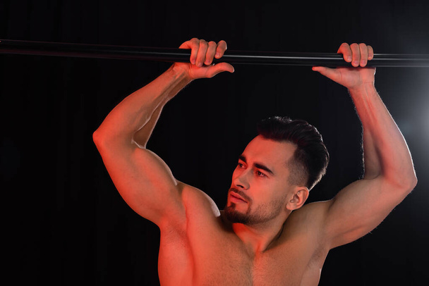 red lighting on muscular sportsman looking away and leaning on horizontal bar isolated on black  - Photo, Image
