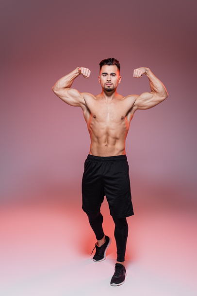 full length of strong man showing muscles and posing on pink and grey   - Фото, зображення