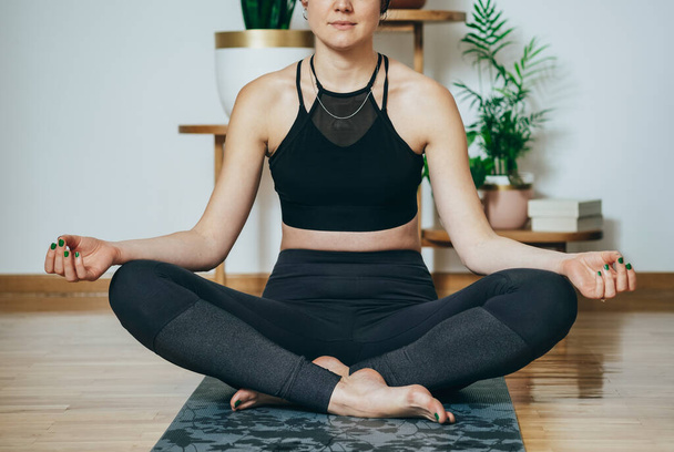 Una donna anonima che pratica yoga al mattino. Donna irriconoscibile in abbigliamento sportivo nero seduta in posizione loto e meditando a casa.  - Foto, immagini