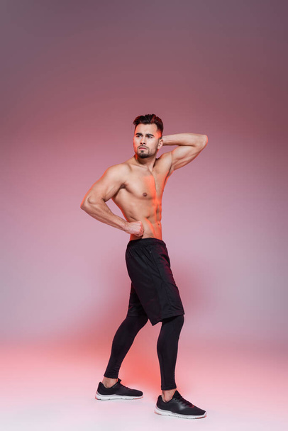 full length of shirtless sportsman posing while showing muscles on grey and pink  - Φωτογραφία, εικόνα