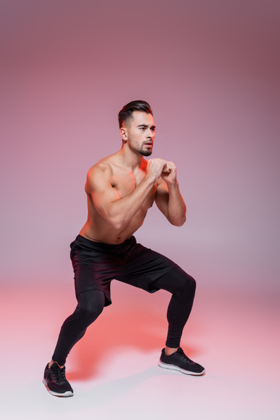 full length of shirtless sportsman doing squat exercise on grey and pink  - Foto, Imagem
