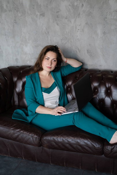 distant work, remote study. confident brunette woman in green suit barefoot on leather couch with laptop. home office. work from home - Foto, Bild