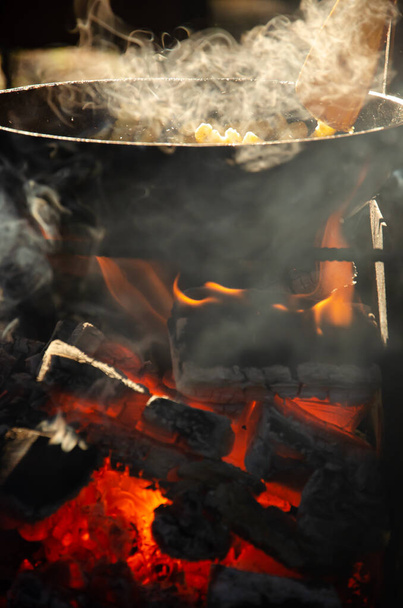 A comida é frita em panela sobre fogo aberto na grelha. O processo de cozinhar sobre o fogo. Foto vertical da comida. Fumaça sobre a comida em chamas no fundo da floresta. - Foto, Imagem