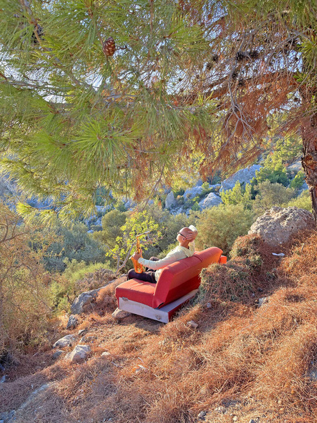 Mladý stylový plešatý saxofonista sedí na pomerančové pohovce v horách Kypru v přírodě, pod borovicí, drží v rukou alt saxofon. Hudebník se saxofonem na přírodě v horách. - Fotografie, Obrázek