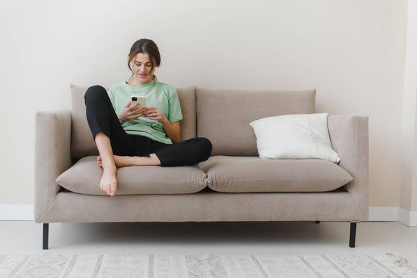 Imagen de una joven feliz y optimista sentada en casa y comprando o en las redes sociales usando el teléfono móvil en el sofá en casa. - Foto, Imagen