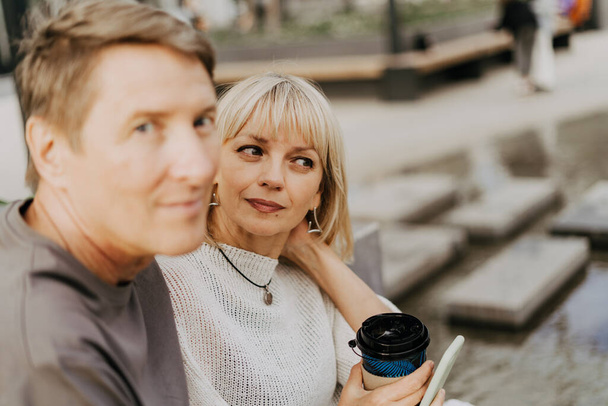 An adult mature happy couple in love sitting on bench outdoors in city street park. A blonde caucasian man and woman spend time together and drinking coffee. Senior wife and husband walking outside. - Photo, Image
