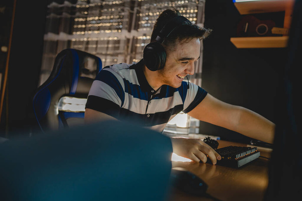 Jeden mężczyzna młody biały mężczyzna gra w gry wideo na komputerze stacjonarnym w domu w ciemnym pokoju sam ze słuchawkami na głowie szczęśliwy uśmiechnięty prawdziwi ludzie kopiować przestrzeń - Zdjęcie, obraz