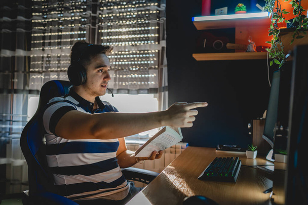 Jeden młody człowiek kaukaski mając zajęcia online, siedząc w ciemnym pokoju przy komputerze stacjonarnym PC trzymając książkę podczas lekcji Internetu jako mentor lub nauczyciel freelance zdalnej edukacji prawdziwych ludzi - Zdjęcie, obraz