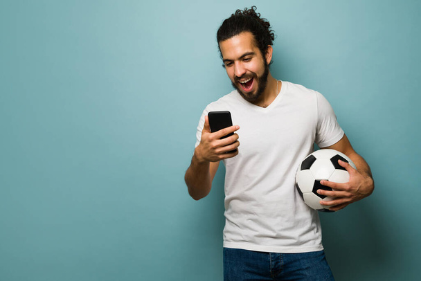 Aficionado al fútbol latino leyendo noticias en línea de su equipo favorito. Joven hombre mensajes de texto a sus amigos para jugar fútbol - Foto, imagen