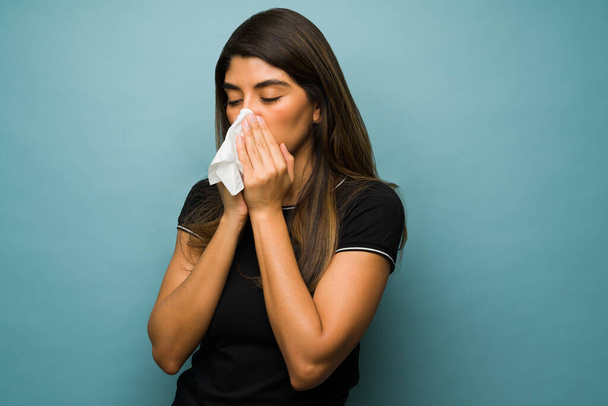 Runny nose. Sick hispanic woman blowing her nose with a tissue and suffering from a bad cold - Photo, Image