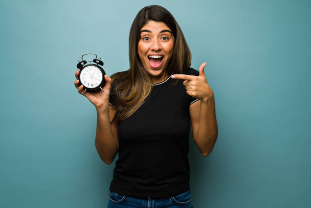 Daylight saving time. Beautiful hispanic woman showing an alarm clock and looking happy about the time change - Photo, Image