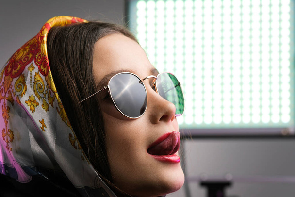Attractive sensual woman posing in studio with a lamp for camera in beautiful headscarf and sunglasses showing her tongue. - Foto, Imagem