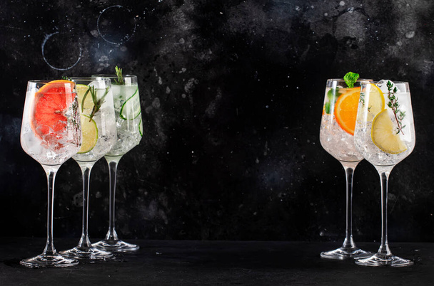Gin and tonic cocktails set. Trendy Alcoholic drinks with lime, lemon, grapefruit, orange, cucumber, soda and spicy herbs in wine glasses, black background. Summer cocktail party - Foto, Imagem