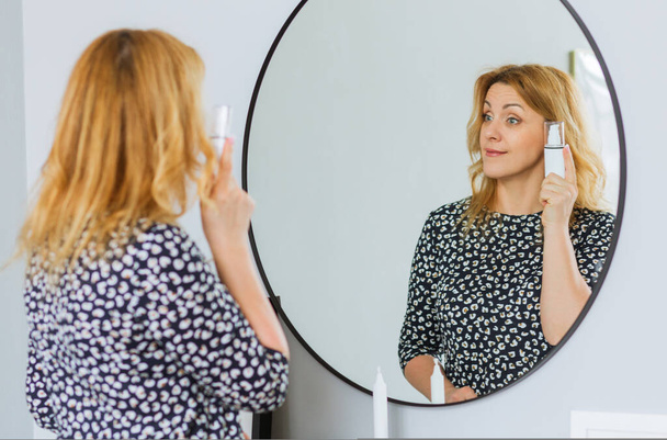 Une belle femme mûre dans une robe à la mode se tient devant un miroir et tient une crème pour le visage dans ses mains. Concept d'autosoin après 40 ans - Photo, image