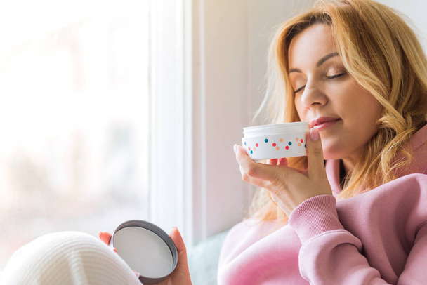 A mature woman in a pink hoodie sits on the windowsill, holds a jar of cream in her hands and sniffs it. The concept of a beautiful and healthy life - Photo, Image