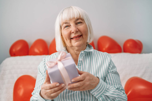 Happy 50 years old blonde woman lady giving gift box on the camera on 14 февраля sitting on a sofa at home. - Фото, изображение