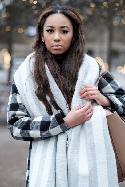 Egy fiatal fekete nő portréja, aki télen a kamerába néz. Kabátot, sálat és táskát visel.. - Fotó, kép