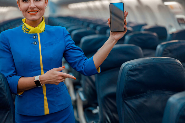 Hymyilevä lentoemäntä kanssa matkapuhelin seisoo lentokoneen matkustamossa - Valokuva, kuva