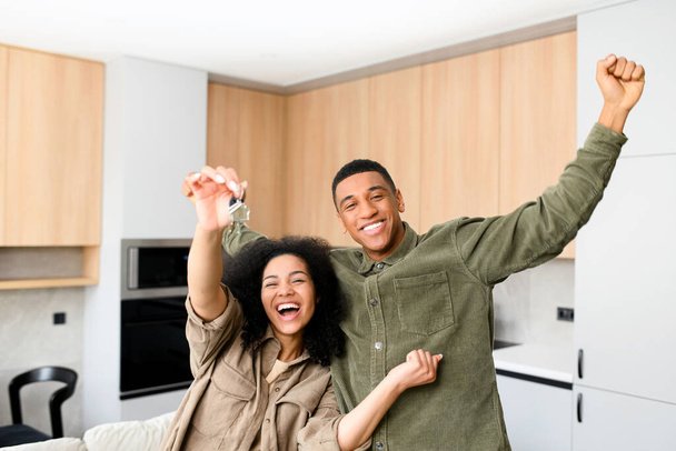 Multiracial couple in love hugging in a new home while holding keys in hands. Young multiracial family bought a new house, rent an apartment - Фото, изображение
