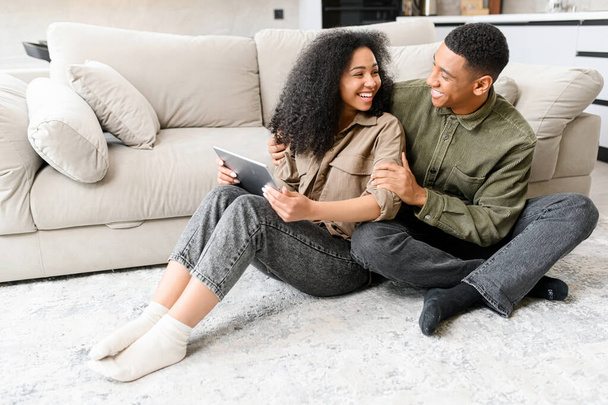 Multiracial girl wearing casual clothes is looking at her husband while sitting together at the floor with tablet - Photo, Image