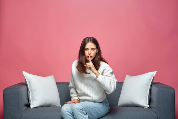 Shh, dont speak. Serious modern girl show silence gesture, ask to stay quiet, sitting on sofa on pink studio background - Foto, Imagen