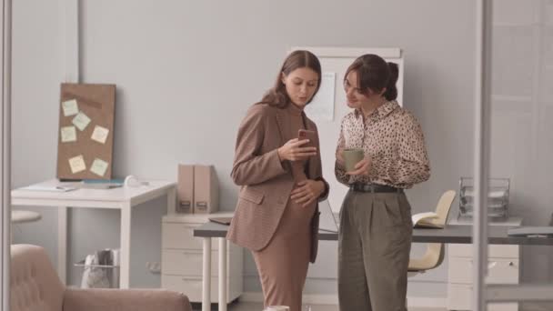 Keskipitkä hidas kuva nuoresta valkoihoisesta raskaana olevasta naisesta, joka näyttää vauvan ultraäänikuvaa älypuhelimella kollegalleen, seisoo työpaikalla kahvitauon aikana - Materiaali, video