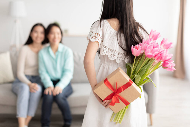 Празднование Дня женщин. Азиатская девушка прячет за спиной подарок и цветы, поздравляя маму и бабушку с праздником - Фото, изображение