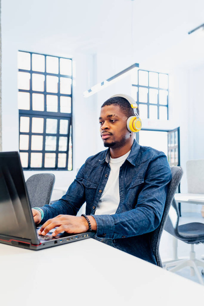 African man with yellow headphones working with a laptop in a coworking office. Black college boy studying and doing work for the university. - 写真・画像
