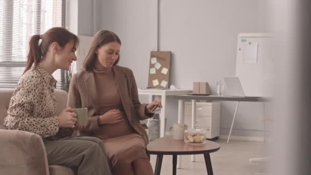 Keskipitkä hidas kuva nuoresta valkoihoisesta raskaana olevasta naisesta, joka näyttää vauvan ultraäänikuvan naispuoliselle kollegalleen, jutteleminen kahvitauon aikana toimistossa - Materiaali, video