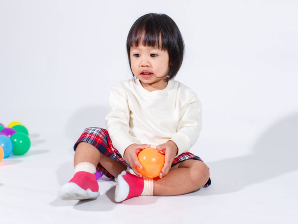 Studioaufnahme von kleinen niedlichen kurzen schwarzen Haaren asiatisches Baby Mädchen Tochter Modell in lässig karierten Rock sitzt auf dem Boden lächelnd lachend mit bunten runden Kugeln Spielzeug allein auf weißem Hintergrund spielen. - Foto, Bild