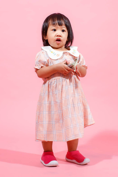 Portrait studio shot little cute Asian kindergarten preschooler girl daughter model in casual long dress and shoes standing look at camera holding hundred dollars banknote in hands on pink background. - Фото, изображение