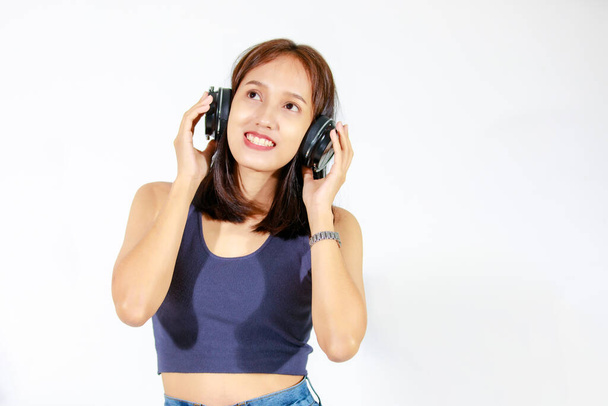 Portret odizolowany studio wycinanka strzał Azji młoda kobieta model w koszuli i dżinsy trzymając duży bezprzewodowy słuchawki stoi uśmiechnięty spojrzenie na aparat słuchać piosenek muzycznych na białym tle. - Zdjęcie, obraz