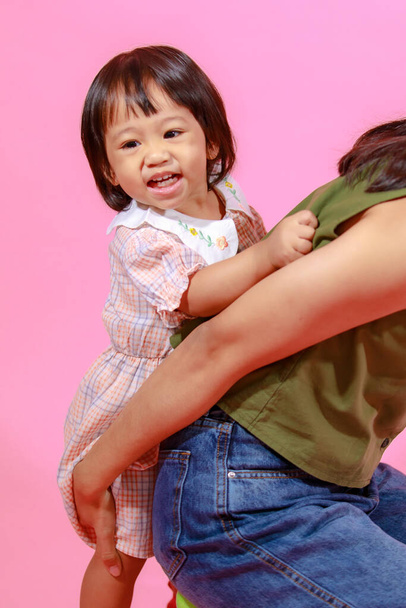 Портрет крупным планом студии снимок азиатской молодой счастливой матери-подростка, держащей обнимашки с маленькой милой дошкольной дочкой, маленькой девочкой, улыбающейся, смотрящей в камеру вместе на розовом фоне. - Фото, изображение