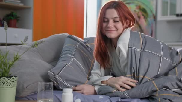 glückliche Frau nimmt Tabletten oder Vitamine und trinkt sie mit kühlem, sauberem Wasser, nachdem sie aus dem Schlaf aufgewacht ist, im Bett liegend, lächelnd und in die Kamera blickend - Filmmaterial, Video