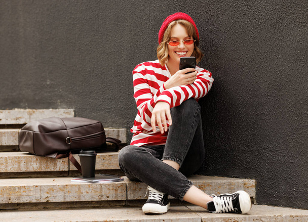 Mosolygó diáklány mobiltelefont használ, miközben kint ül egy kávéval reggel előtt - Fotó, kép