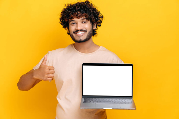 Izgalmas, vidám indiai vagy arab fickó alap pólóban, nyitott laptopot tart üres fehér képernyővel, hüvelykujjával felfelé mutató gesztust mutat, elszigetelt narancssárga háttéren áll, barátságos mosollyal. A modell koncepciója - Fotó, kép